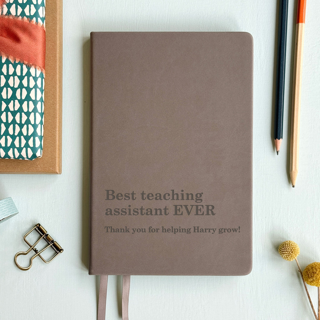 Grey teaching assistant gift notebook engraved best teaching assistant ever text, atop pale background with assorted stationary surrounding.