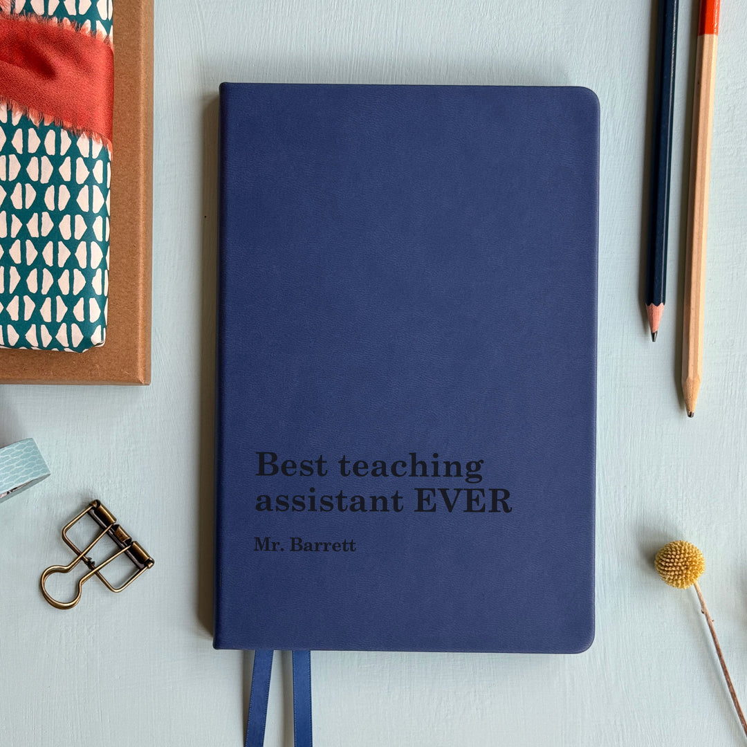 Mid blue Navy teaching assistant gift notebook engraved best teaching assistant ever text, atop pale grey background with assorted stationary surrounding.