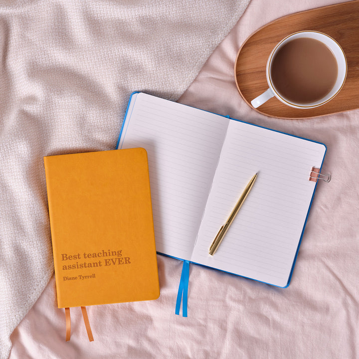 Mustard yellow teaching assistant gift notebook engraved best teaching assistant ever text, atop pale blue open lined notebook atop pink bedsheet background with coffee cup on tray.