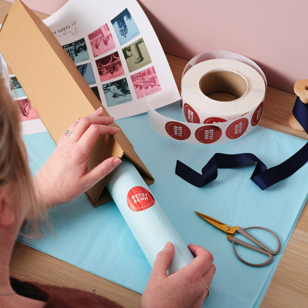 A person packing a rolled-up print into a triangular cardboard box, sealing it with a custom "Betsy Benn" sticker, with packaging materials like ribbon, scissors, and sticker rolls nearby.