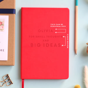 A top down image of a red vegan leather notebook resting on a pale aqua table. The front of the notebook features the phrase "For small thoughts and big ideas". It is personalised with the name Olivia engraved above the text. Surrounding the book are various stationary items and dried flowers. The image is edited to show the area that can be personalised in white text.