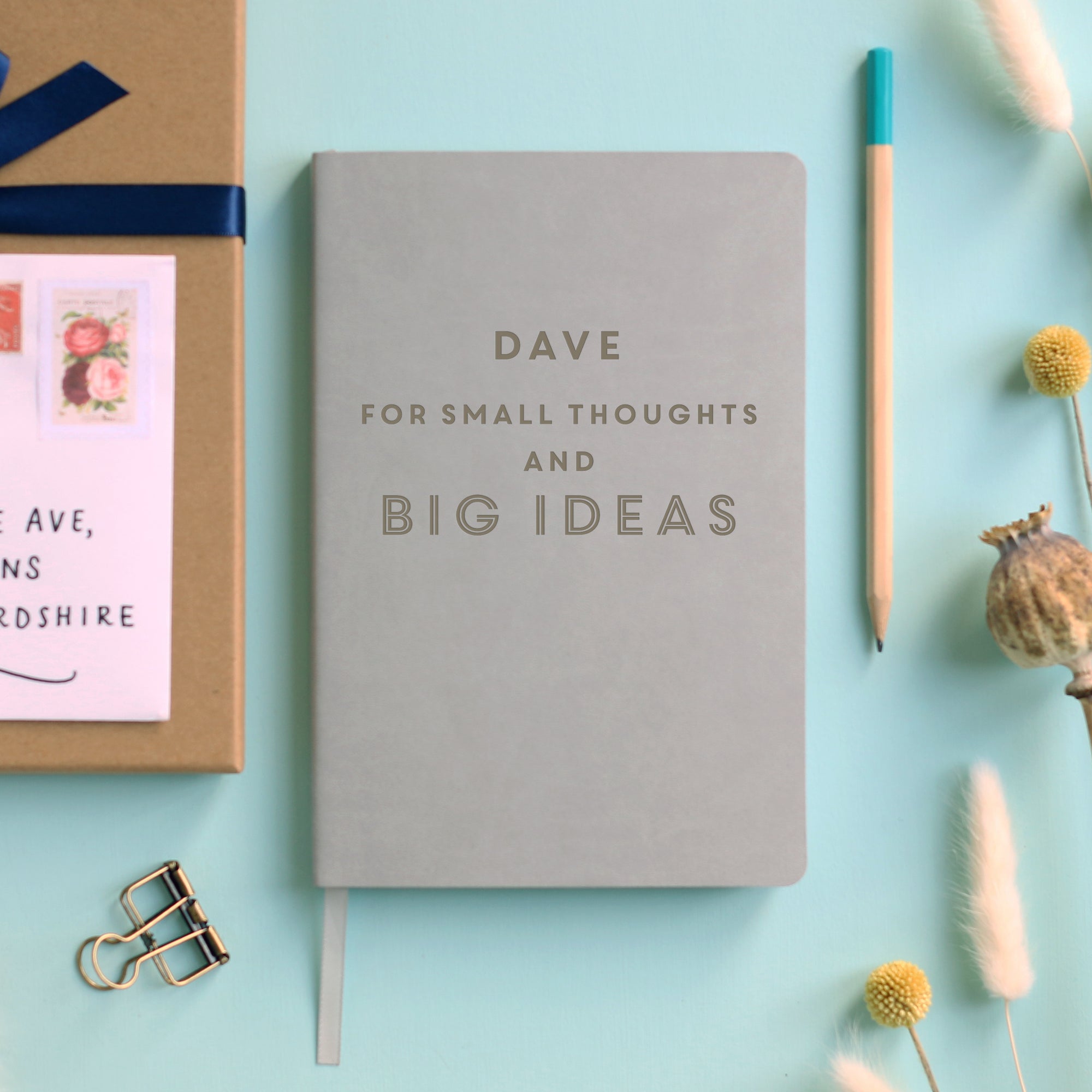 A top down image of a grey vegan leather notebook resting on a pale aqua table. The front of the notebook features the phrase "For small thoughts and big ideas". It is personalised with the name Dave engraved above the text. Surrounding the book are various stationary items and dried flowers.