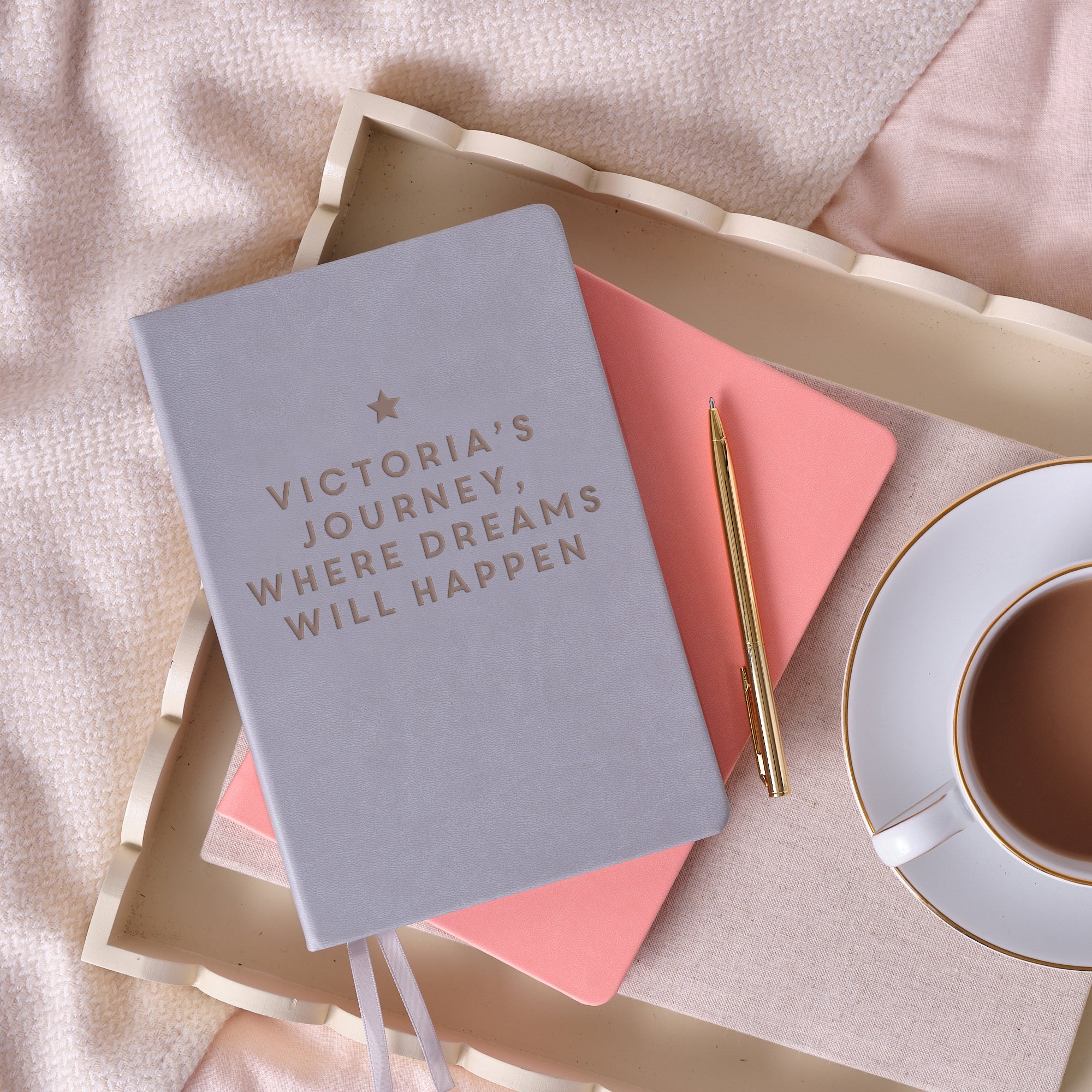 A top down image of a grey vegan leather notebook with the personalised text "Victoria’s journey, where dreams will happen” with a star engraved on it. It is stacked above a pink notebook and is resting on a cream wooden tray with a cup of tea, and a gold pen beside it. The tray is resting on a pink bedspread.