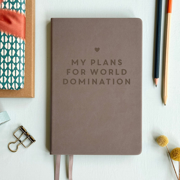 A top down image of a grey vegan leather notebook resting on a pale aqua table. The front of the notebook is engraved with a star and the phrase FOR PLANS AND WORLD DOMINATION in large block text. Surrounding the book are various stationary items.