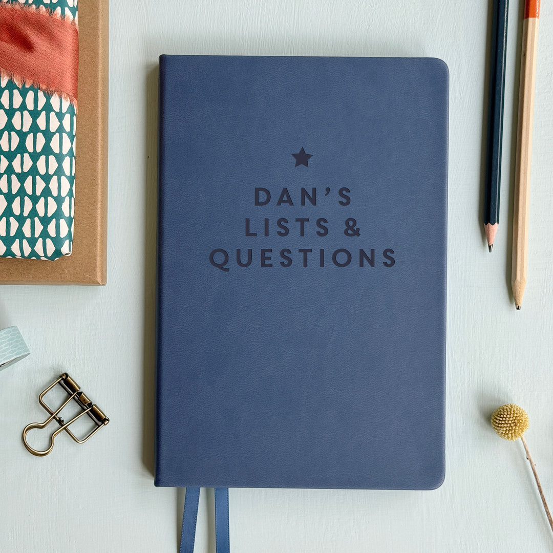 A top down image of a mid blue vegan leather notebook resting on a pale aqua table. The front of the notebook is engraved with a star and the phrase Dan's Lists & Questions in large block text. Surrounding the book are various stationary items.