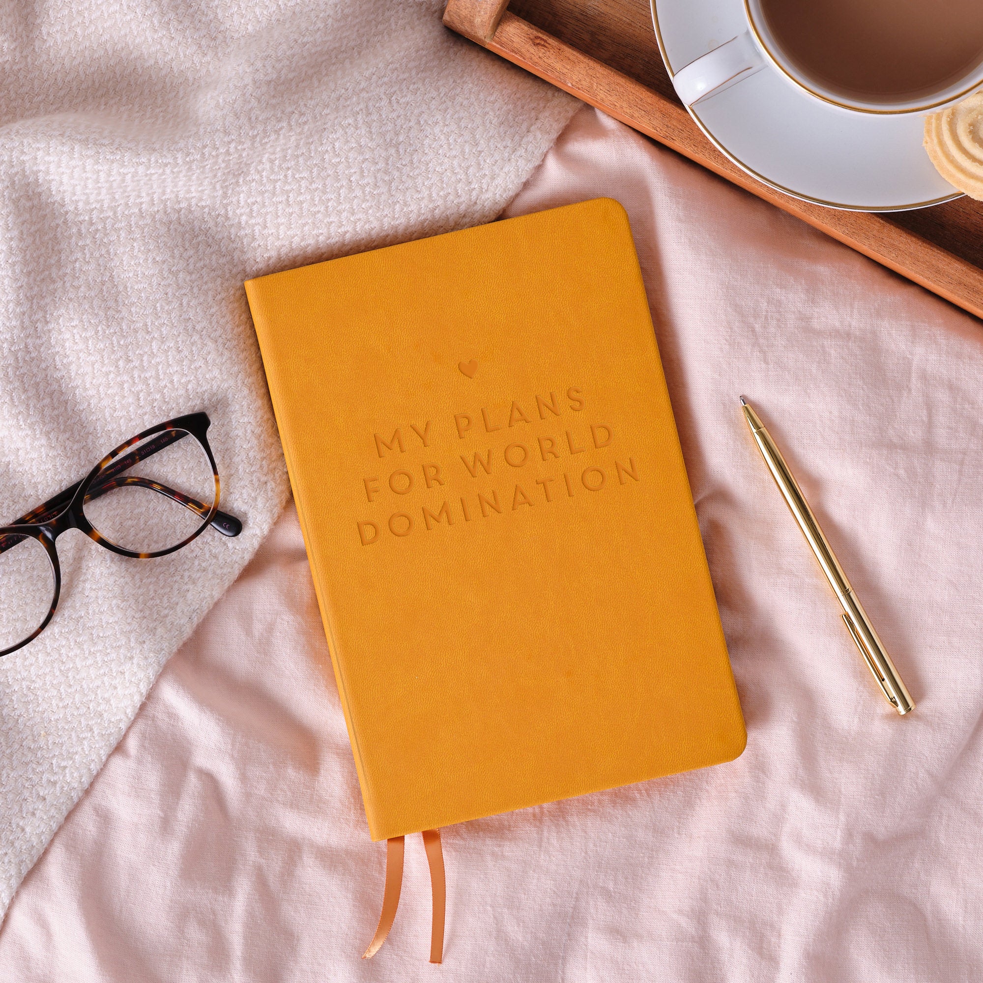 A top down image of a mustard yellow vegan leather notebook resting on a pale pink bed spread. The front of the notebook features personalised engraved text which reads “My plans for world domination” with a heart above it.  Surrounding the book is a tray with a cup of tea and a biscuit, a pair of glasses and a gold pen.