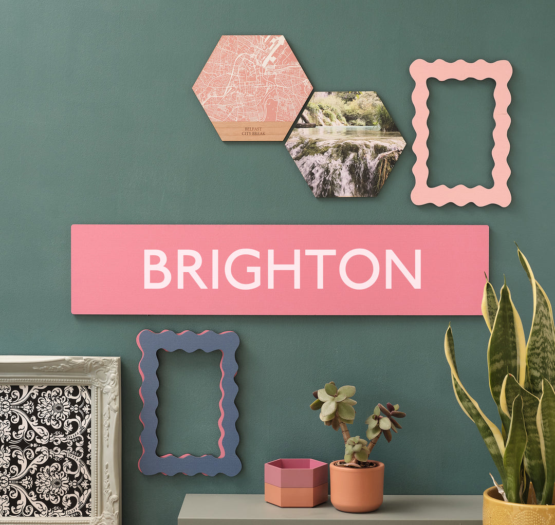 Funky hot pink quirky Brighton wooden wall art sustainable wood sign for those who love their location with pride or wish to celebrate a favourite holiday travel destination. On a forest green wall with hexagonal wooden tiles, picture frames and plants.