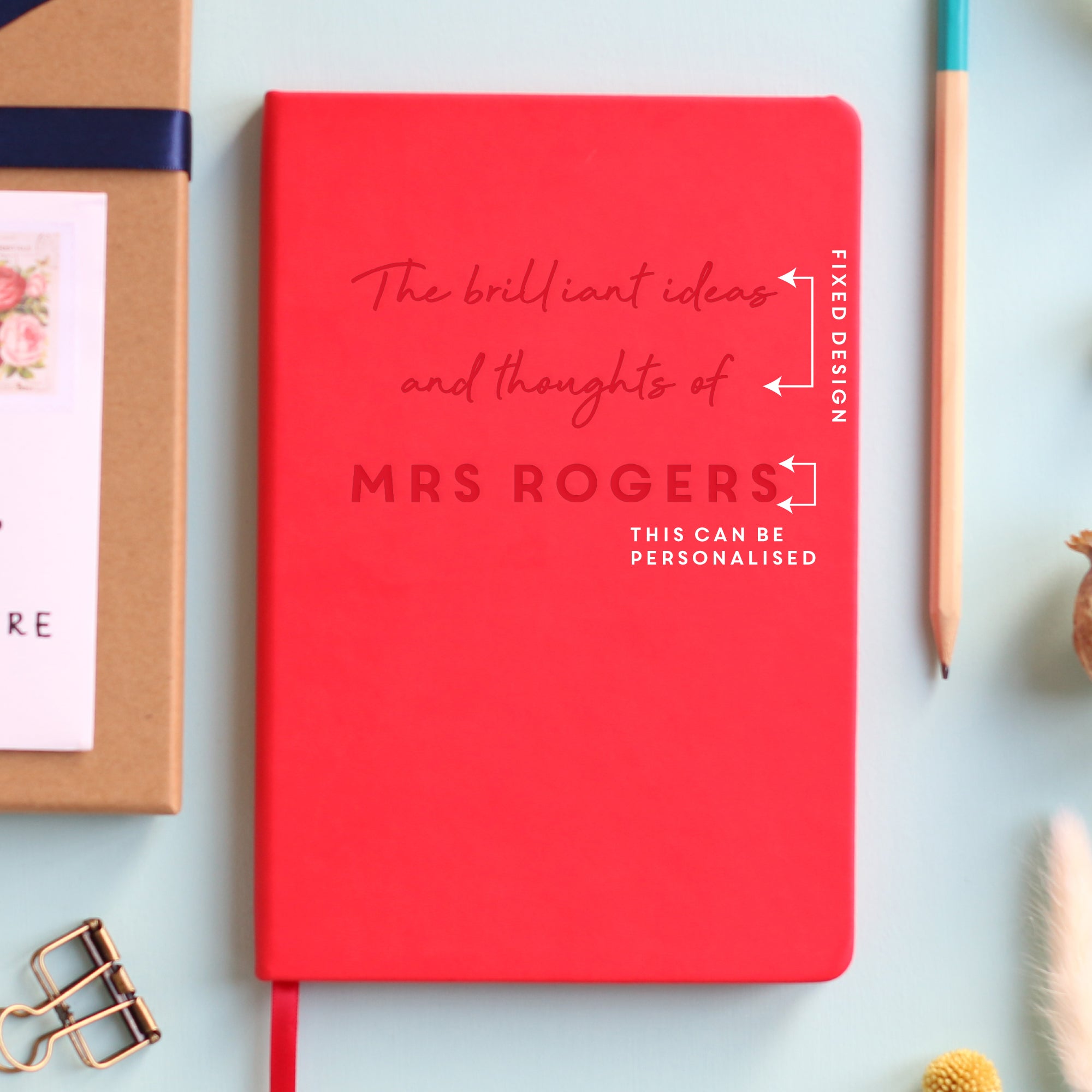 A top down image of a red vegan leather notebook resting on a pale aqua table. The front of the notebook is engraved with the phrase "The brilliant ideas and thoughts of" and is personalised with the name Mrs Rogers. White text highlights what words can be personalised. Surrounding the book are various stationary items and dried flowers.