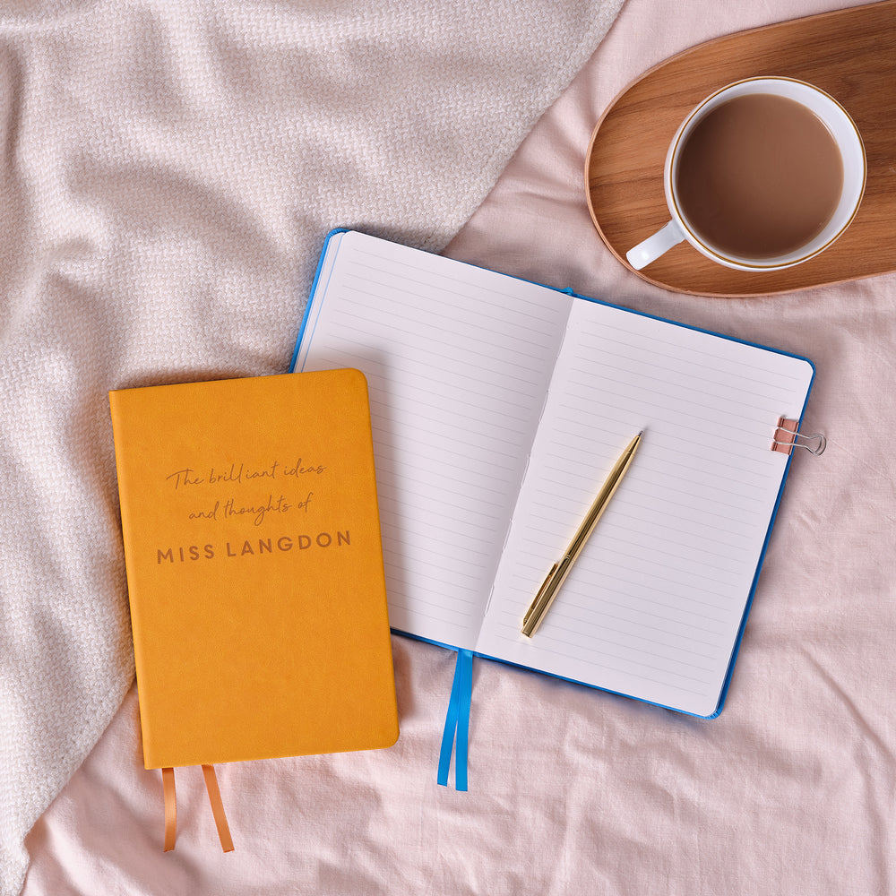 A top down image of a mustard yellow vegan leather notebook with The brilliant ideas and thoughts of Miss Langdon engraved on the front. It is stacked above a blue open notebook with a gold resting on. Next to the books is a tray with a cup of coffee on. The tray and books are resting on a pink bedspread.