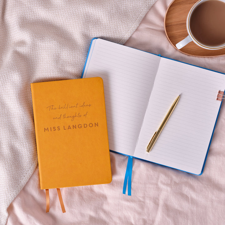 A top down image of a mustard yellow vegan leather notebook with The brilliant ideas and thoughts of Miss Langdon engraved on the front. It is stacked above a blue open notebook with a gold resting on. Next to the books is a tray with a cup of coffee on. The tray and books are resting on a pink bedspread.