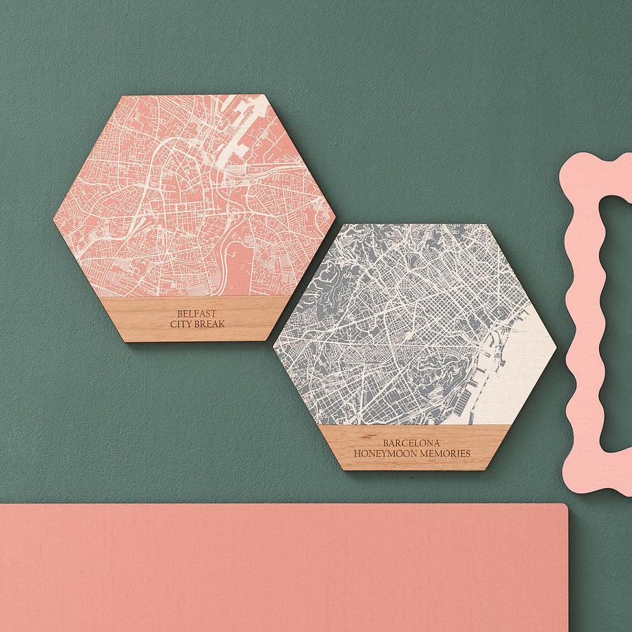 Two hexagon-shaped wooden map prints on a green background, one with a pink map print and the other with a white map print. Both have personalised text at the bottom, indicating a city name from a travel destination. Perfect to remember that special place in an anniversary gift.