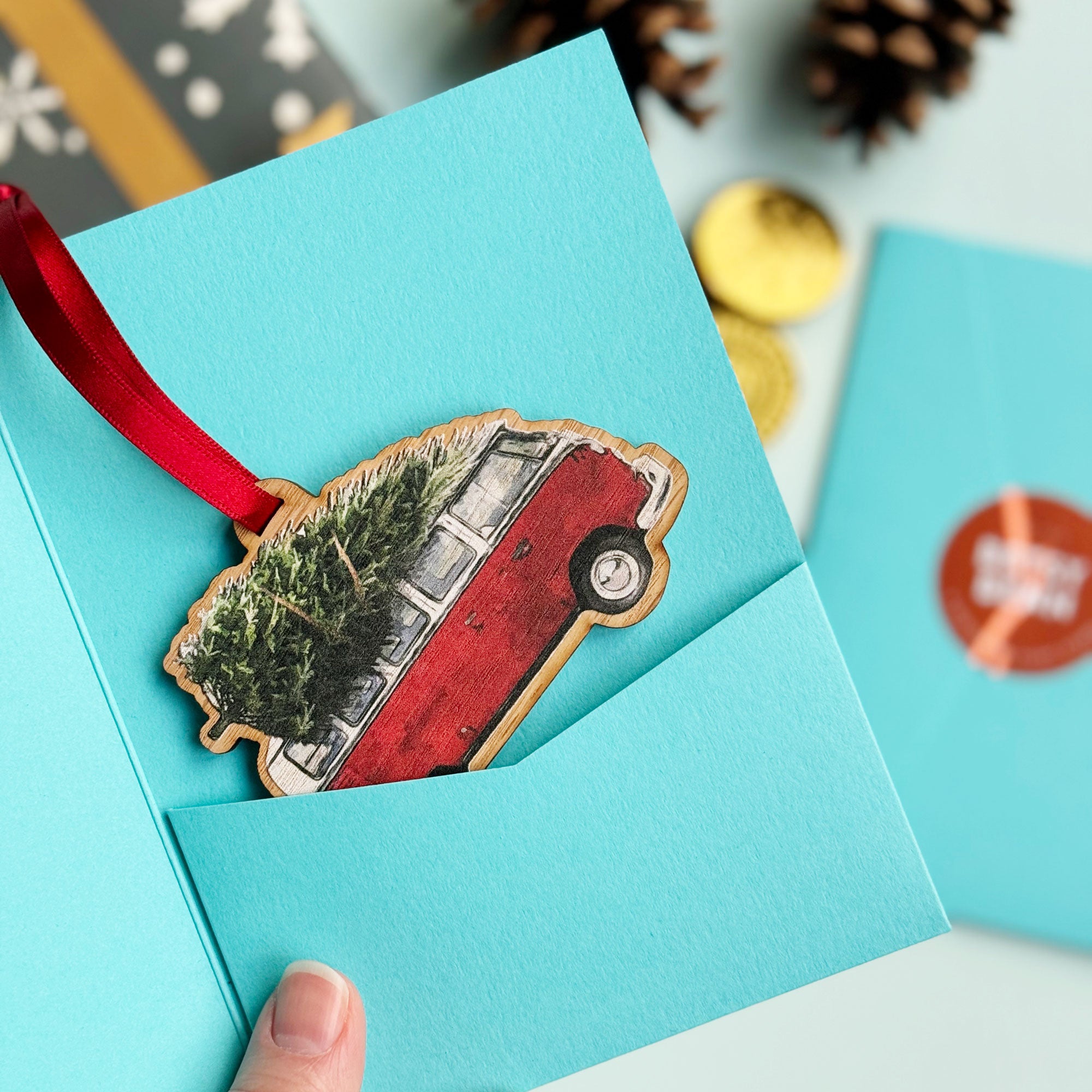  A wooden Christmas ornament shaped like a vintage red VW camper van with a Christmas tree strapped to the roof. It's hanging from a red ribbon on a Christmas tree.