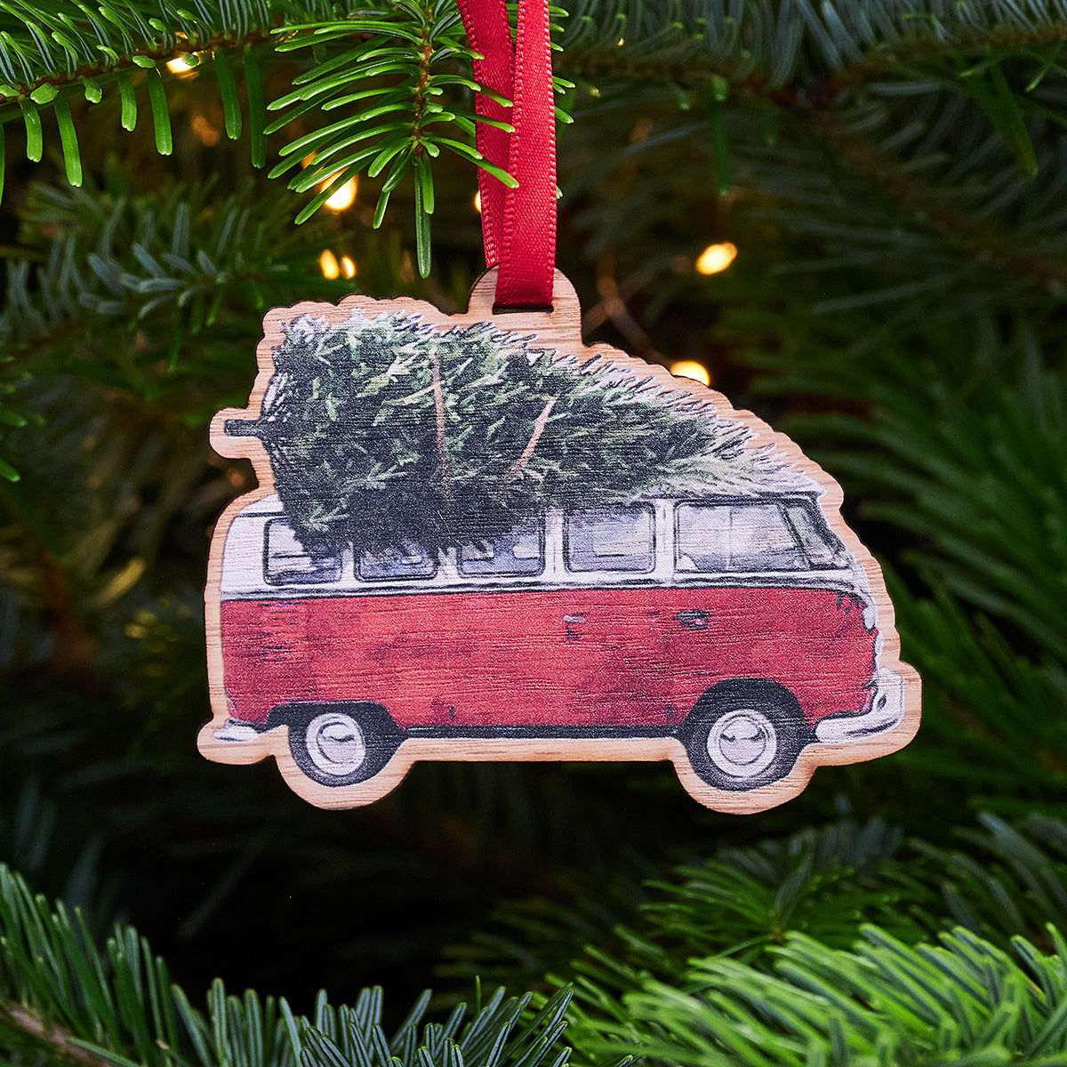  A wooden Christmas ornament shaped like a vintage red VW camper van with a Christmas tree strapped to the roof. It's hanging from a red ribbon on a Christmas tree.