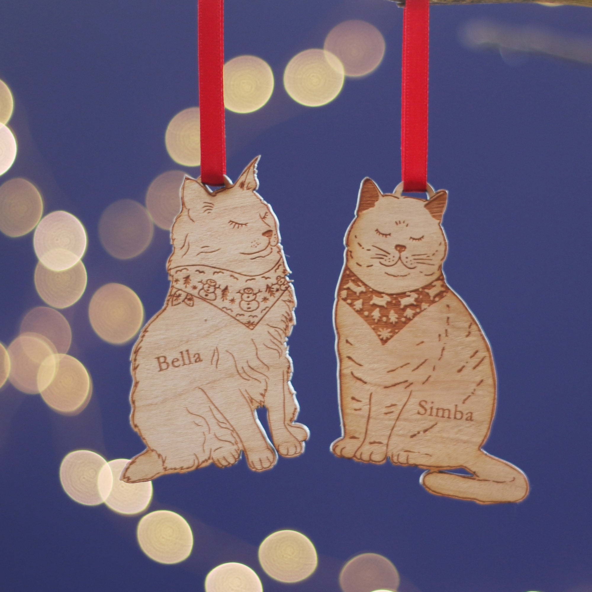 Two cat-shaped wooden christmas tree decorations with custom pet names 'Bella' and 'Simba' on a blue background with bokeh lights. Treat the animal lover in your life to a bespoke pet gift for secret santa or a stocking filler. Would work for British Shorthair, Persian, Maine Coon, Ragdoll cat, and more!