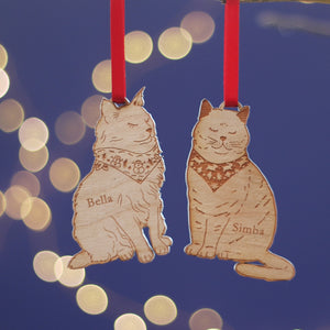 Two cat-shaped wooden christmas tree decorations with custom pet names 'Bella' and 'Simba' on a blue background with bokeh lights. Treat the animal lover in your life to a bespoke pet gift for secret santa or a stocking filler. Would work for British Shorthair, Persian, Maine Coon, Ragdoll cat, and more!