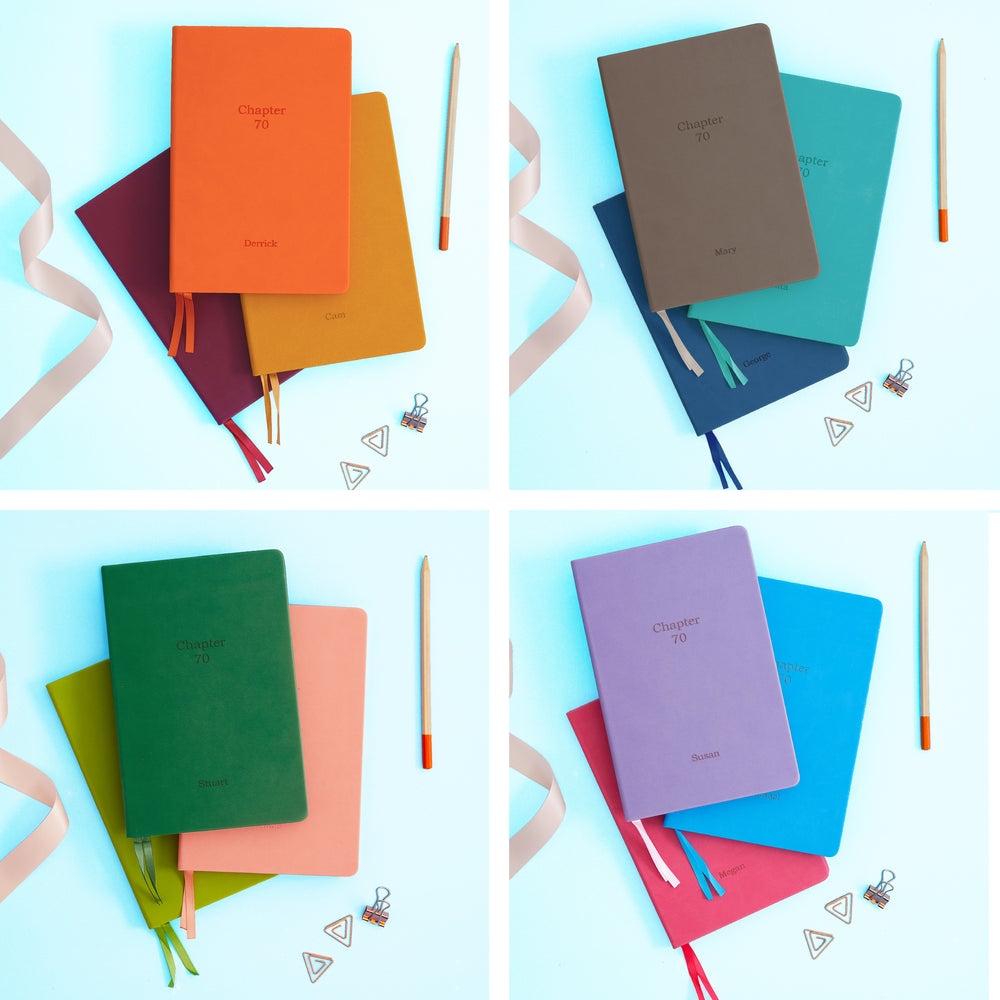 The image shows examples of personalised engraved text on all 12 colours of notebook. The books are stacked in in threes and are surround by various pieces of stationery.