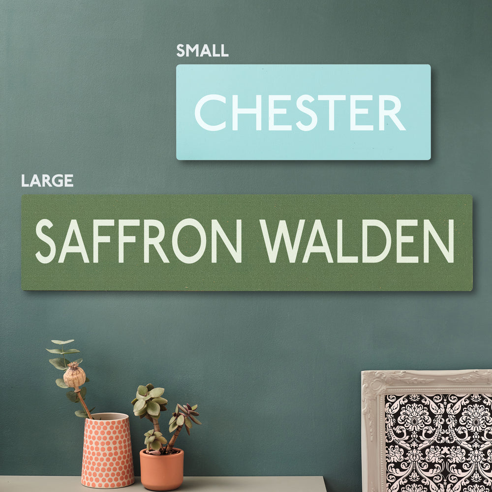 Two rectangular signs with text on a green wall, with decorative items below displaying the size difference between the small and large size wooden sustainable cherry wall art prints. Personalised with colour and location of your choice!