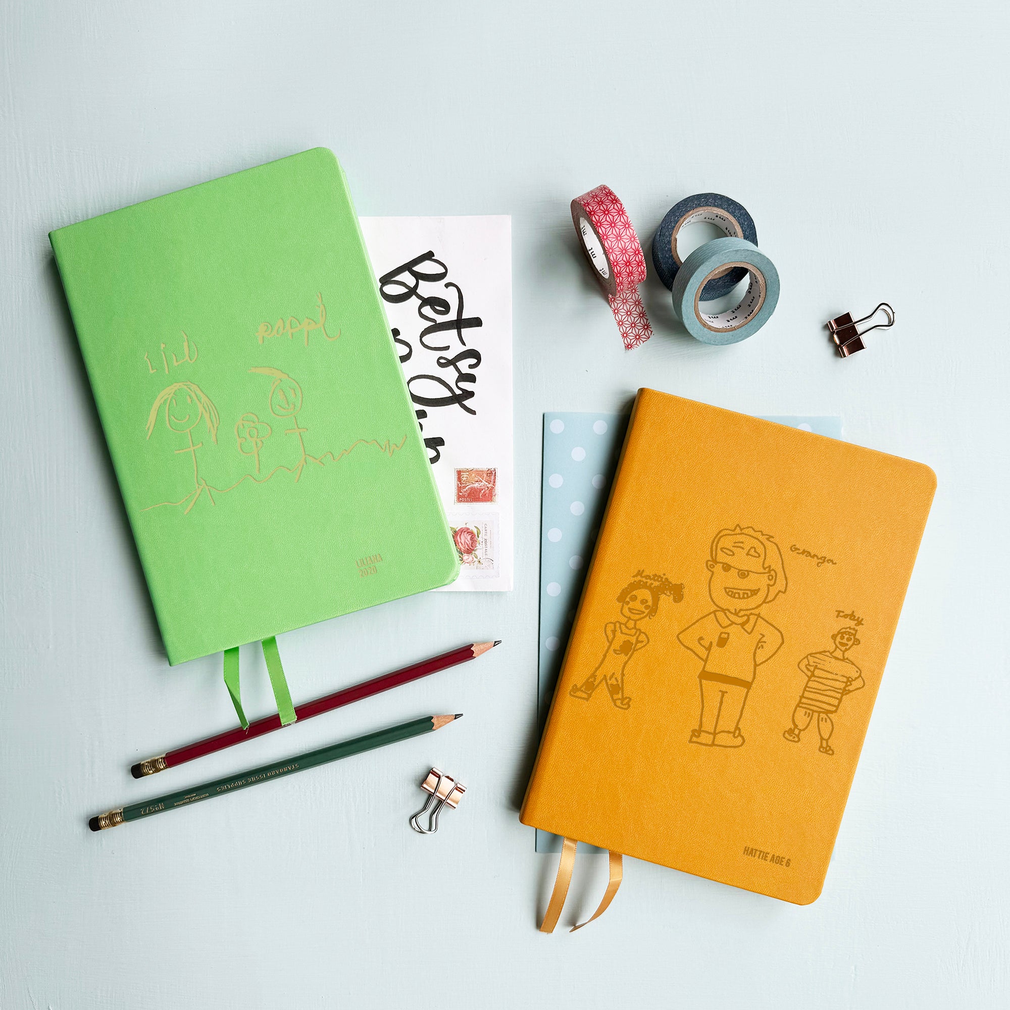 A top down image of two vegan leather notebooks, one green and one mustard yellow. They are resting on a pale aqua table. The front of each notebook is personalised with an engraving of a child’s drawing  Surrounding the book are various stationary items.