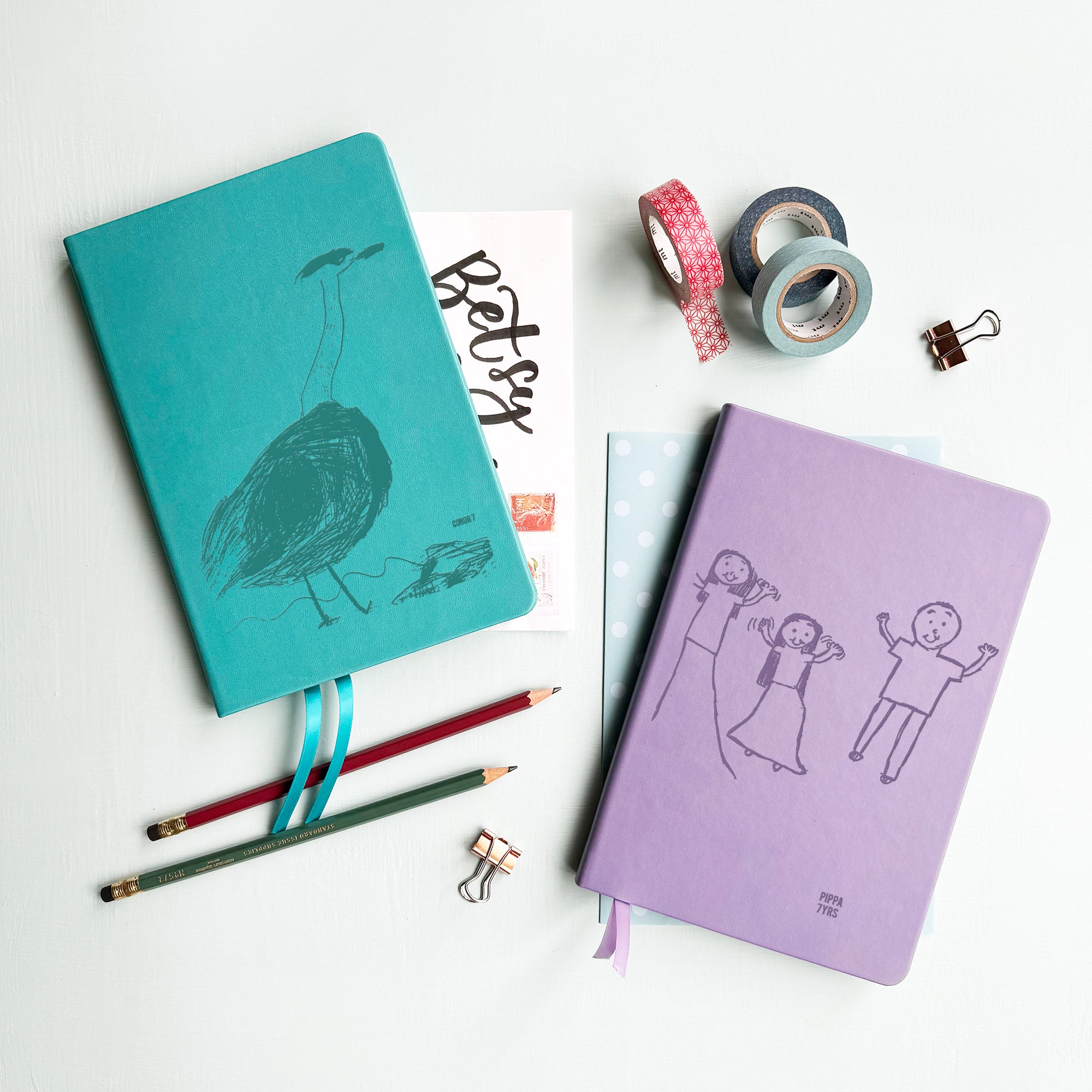 A top down image of two vegan leather notebooks, one turquoise and one lilac. They are resting on a pale aqua table. The front of each notebook is personalised with an engraving of a child’s drawing  Surrounding the book are various stationary items.