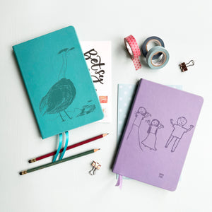 A top down image of two vegan leather notebooks, one turquoise and one lilac. They are resting on a pale aqua table. The front of each notebook is personalised with an engraving of a child’s drawing  Surrounding the book are various stationary items.