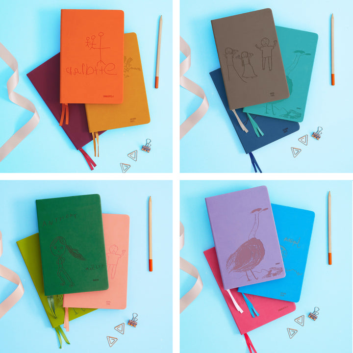 The image shows examples of personalised engraved text on all 12 colours of notebook. The books are stacked in threes and are surrounded by various pieces of stationery.