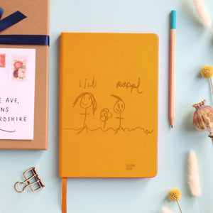 A top down image of a mustard yellow vegan leather notebook resting on a pale aqua table. The front of the notebook is personalised with an engraving of a child's drawing and the text Liliana 2020. Surrounding the book are various stationary items and dried flowers.