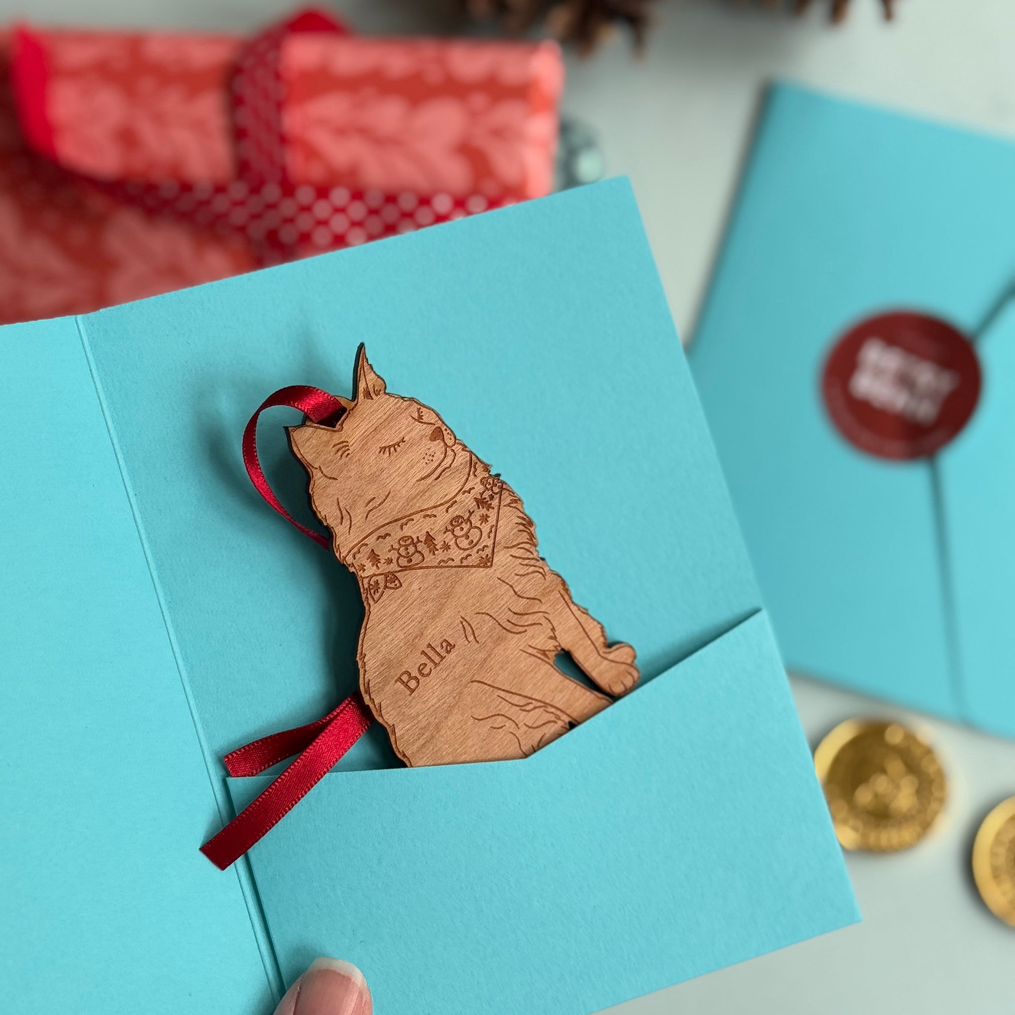 Blue card gift sleeve envelope with a laser-cut wooden cat ornament for a Christmas tree bauble, strung with red ribbon, surrounded by festive gold coins and more cards and presents. Perfect Christmas Gifts for Dog, Cat and Pet Lovers.