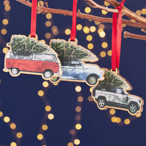 Three wooden Christmas ornaments hanging from a tree branch. Each ornament is shaped like a different vintage car with a Christmas tree strapped to the roof: a red Volkswagen van, a blue Morris Minor, and a Land Rover.