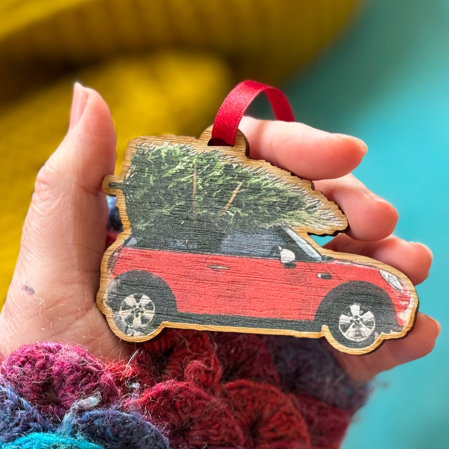 A hand holding a wooden Mini Cooper Christmas tree decoration with a tree on the car roof.