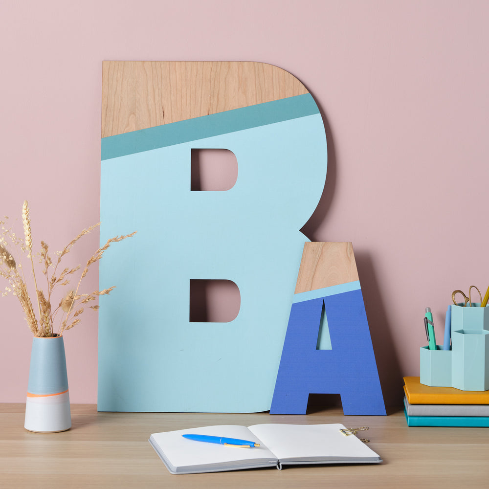 Large wooden letters "B" and "A" painted in shades of blue and teal, displayed on a desk with other stationery items.