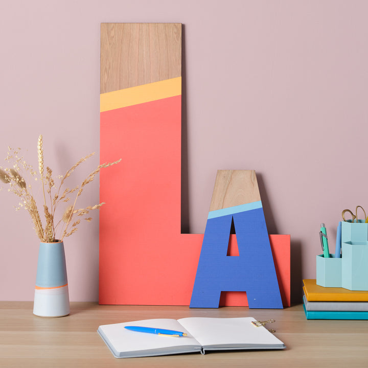 Large wooden letters "L" and "A" painted in vibrant colors.The letters are showcased on a wall above a desk with a vase of dried flowers, stationery items, and a notebook, creating a cheerful and personalized workspace.