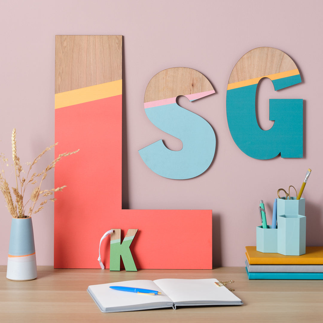 Large wooden letters "L," "S," and "G" painted in vibrant colors, displayed on a wall above a desk with other stationery items.

