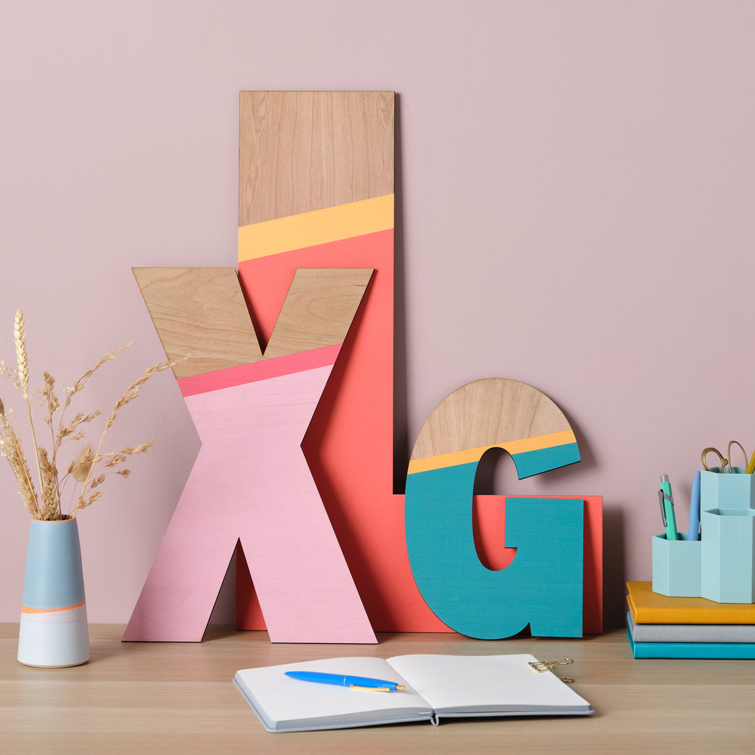 A vibrant and playful display featuring large wooden letters "X", "L" and "G" painted in a striking color palette of pink, orange, and teal, displayed on a desk with other stationery items.