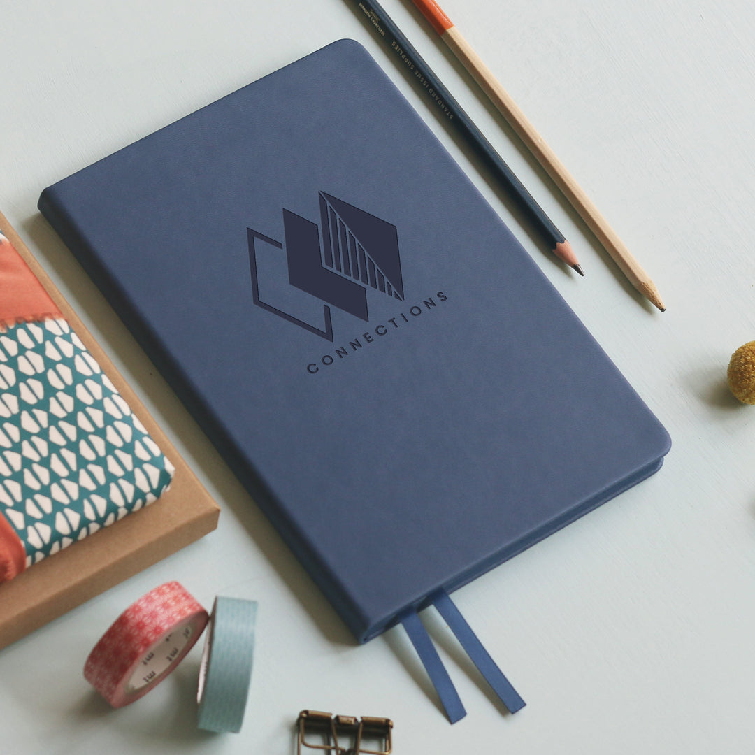 A top down image of a mid blue vegan leather notebook resting on a pale aqua table. The front of the notebook is personalised with an engraving of a business logo. Surrounding the book are various stationary items.