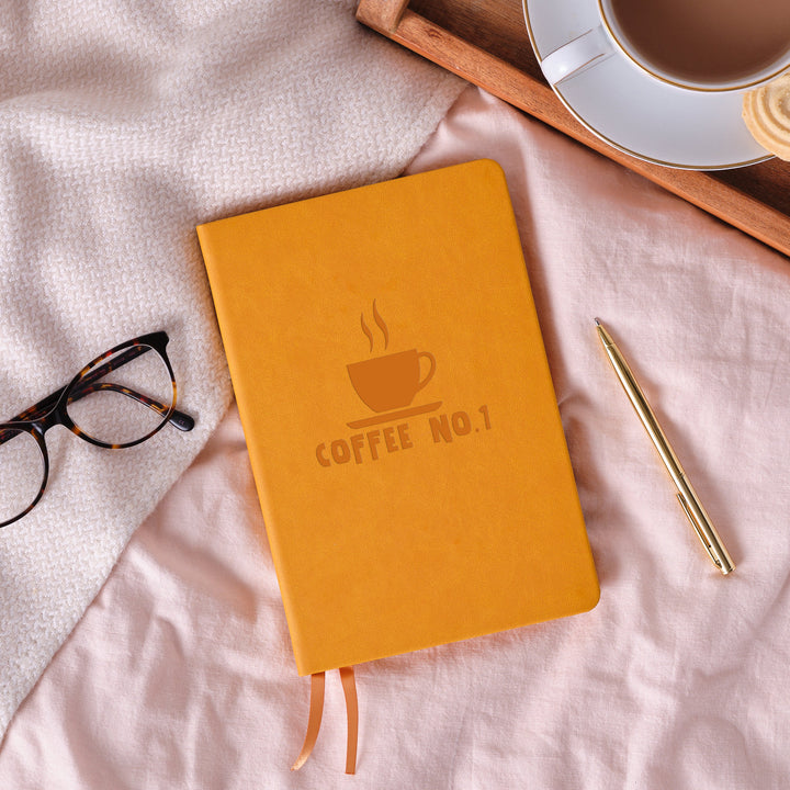 spread. The front of the notebook is personalised with an engraving of a business logo. Surrounding the book is a tray with a cup of tea and a biscuit, a pair of glasses and a gold pen.