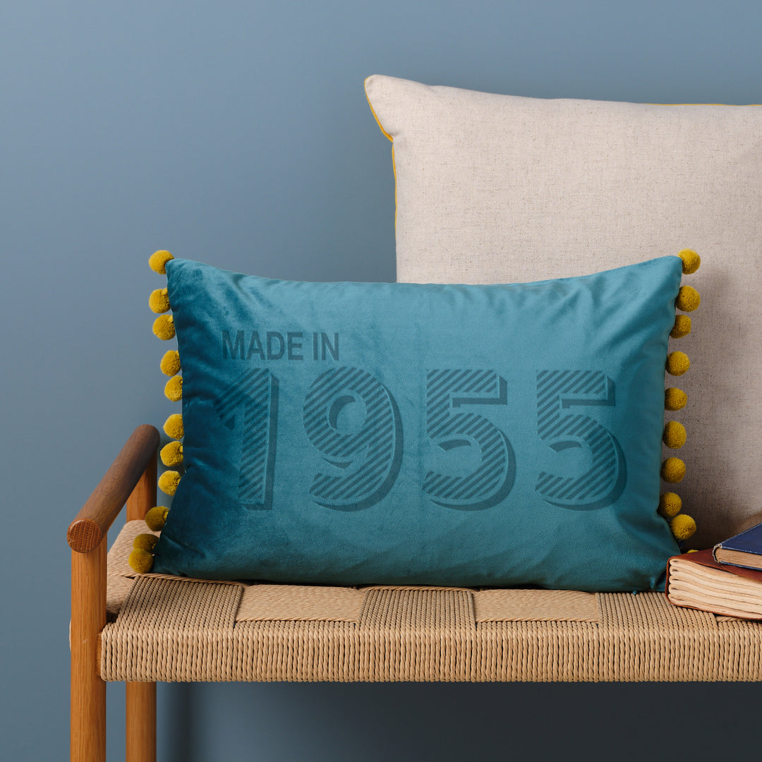 Ocean custom velvet custom cushion personalised with "made in 1955", sitting on a wicker bench in front of a blue wall. Perfect 70th birthday gift for Grandad.