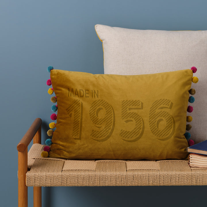 Bamboo/multi custom velvet custom cushion personalised with "made in 1956", sitting on a wicker bench in front of a blue wall. Perfect 70th birthday gift for parents and godparents.
