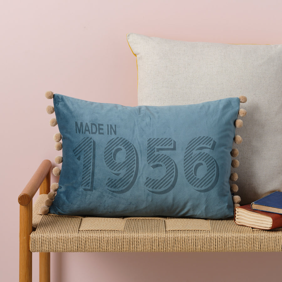 A personalised velvet 70th birthday cushion with 'MADE IN 1956'  engraved on the front, placed on a wicker bench with other cushions and books in the background.