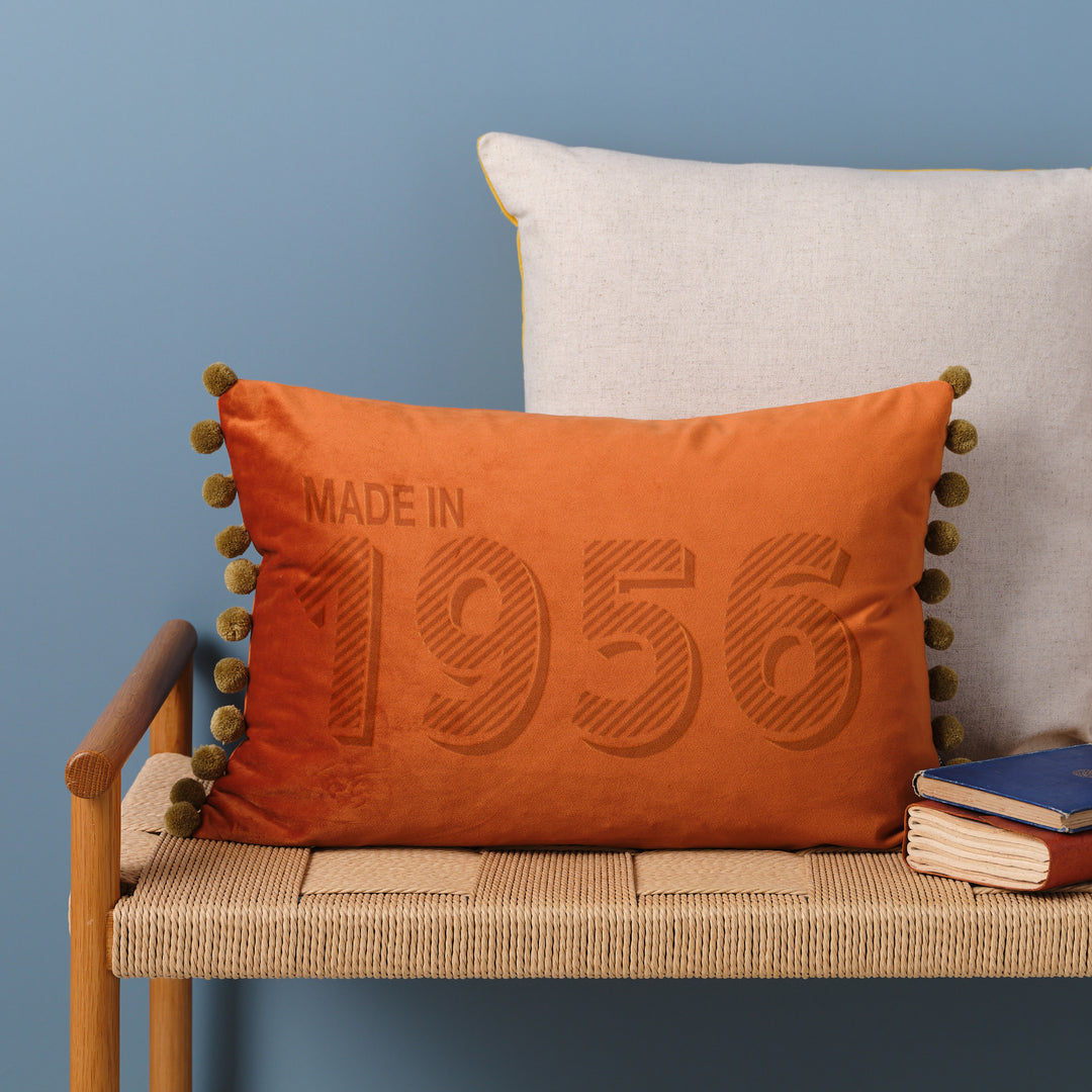 Rust custom velvet custom cushion personalised with "made in 1956", sitting on a wicker bench in front of a blue wall. Perfect 70th birthday gift for Godmothers.