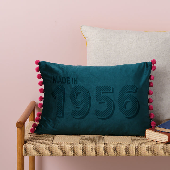 Teal custom velvet custom cushion personalised with "made in 1956", sitting on a wicker bench in front of a blue wall. Perfect 70th birthday gift for mothers day or fathers day.