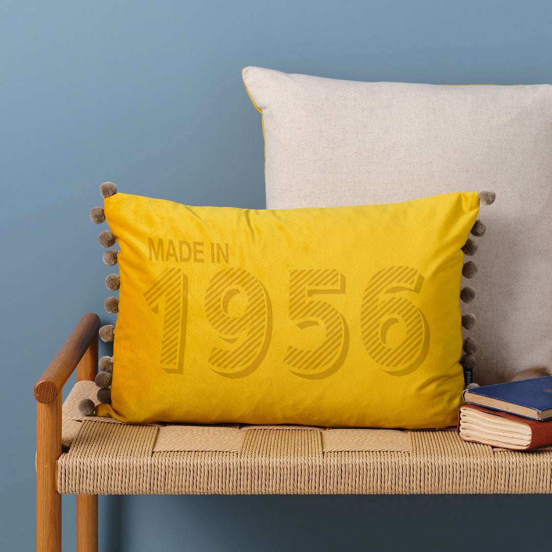 Yellow velvet cushion personalised with "made in 1956", sitting on a wicker bench in front of a blue wall. Perfect 70th birthday gift for Mum or Dad