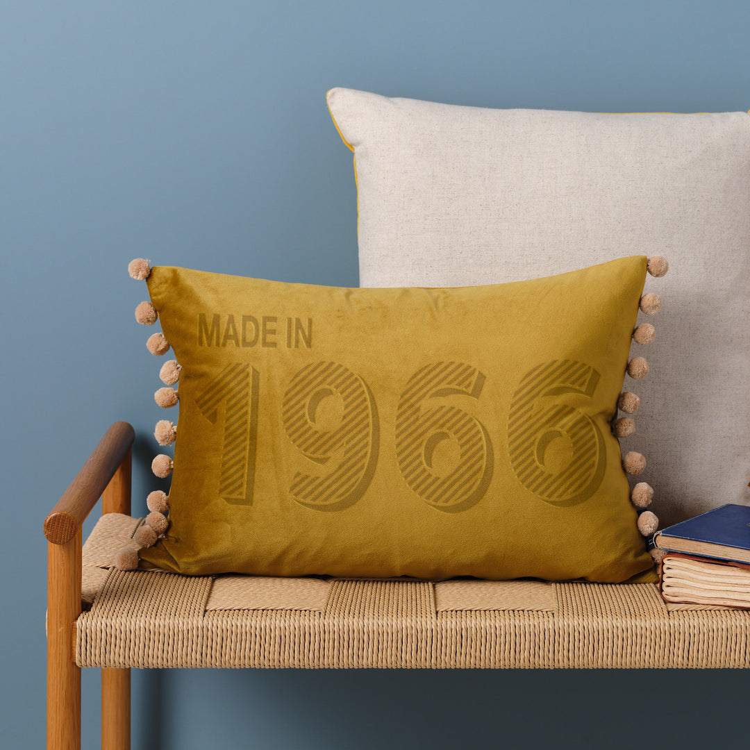 A personalised 60th birthday present for Dad velvet cushion with 'MADE IN 1966' laser engraved on it, placed on a wicker bench with other cushions and a book.