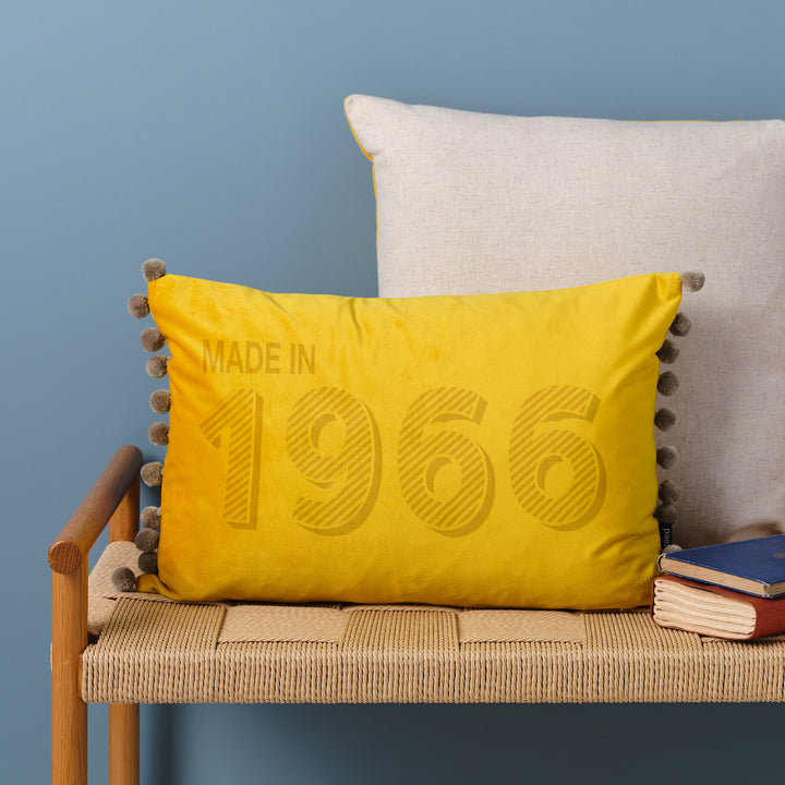 A personalised 60th birthday gift velvet cushion with 'MADE IN 1966' laser engraved on it, placed on a wicker bench with other cushions and a book.