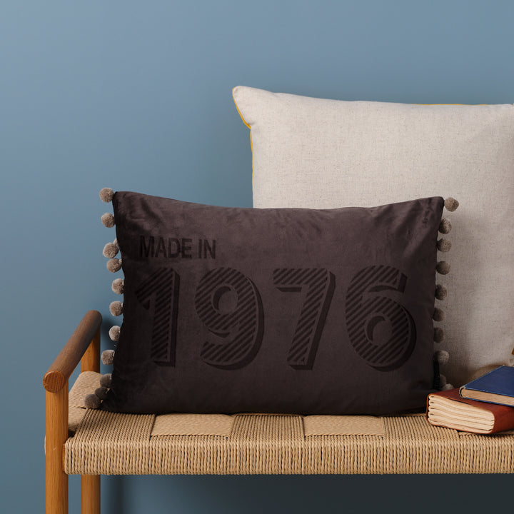 A personalised 50th birthday gift velvet cushion with 'MADE IN 1976' laser engraved on it, placed on a wicker bench with other cushions and a book.