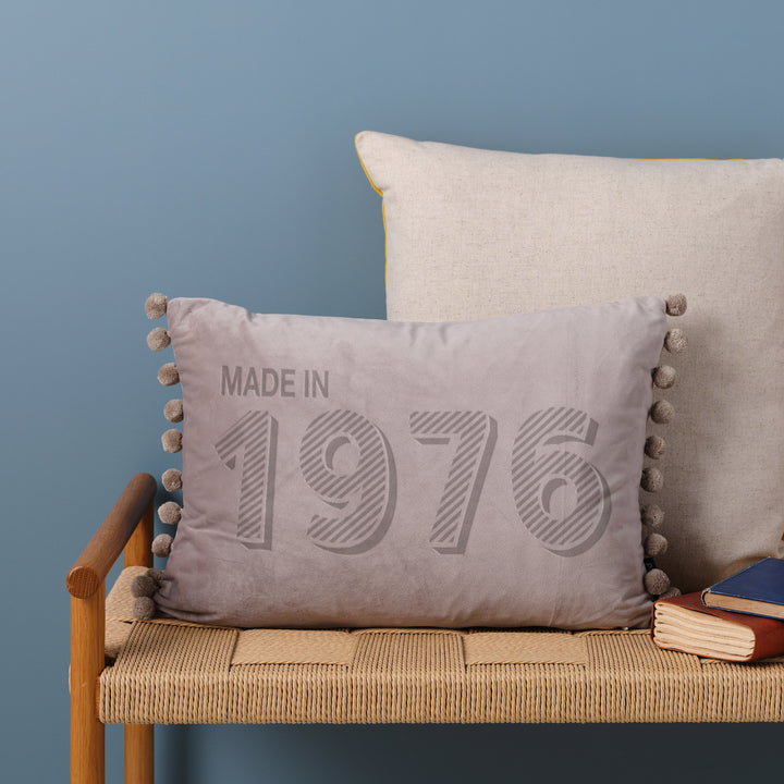 A personalised 50th birthday gift velvet cushion with 'MADE IN 1976' laser engraved on it, placed on a wicker bench with other cushions and a book.
