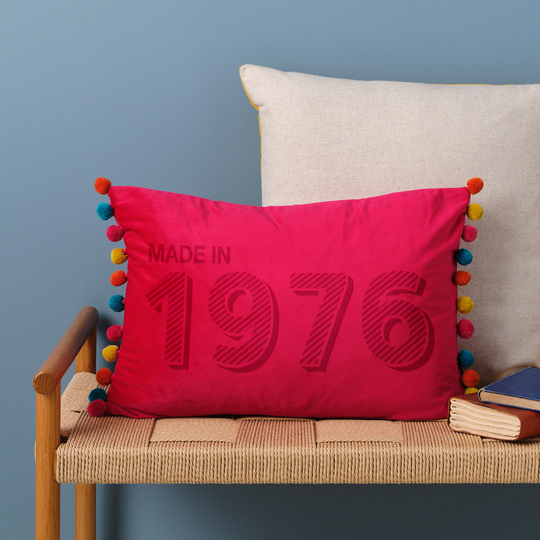 A personalised 50th birthday gift velvet cushion with 'MADE IN 1976' laser engraved on it, placed on a wicker bench with other cushions and a book.