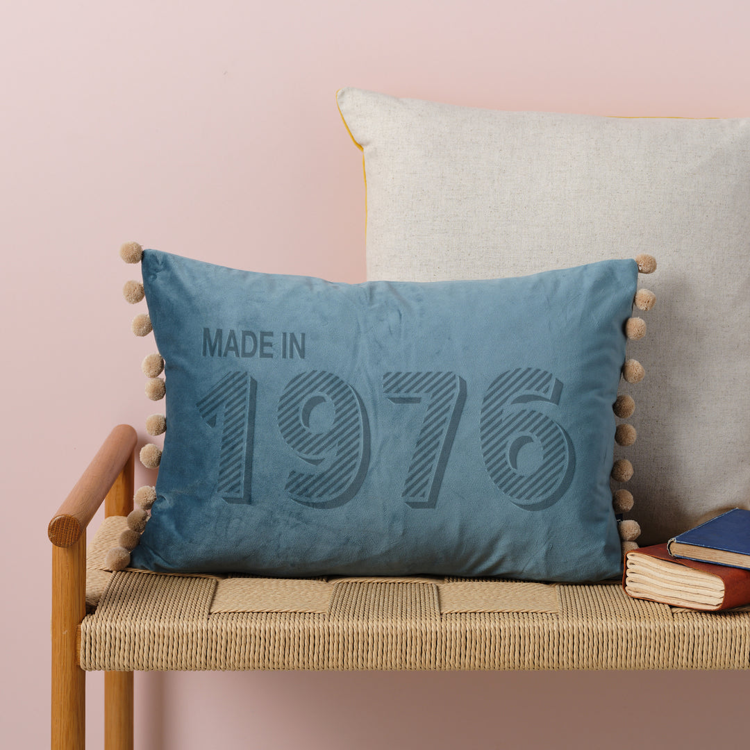A personalised 50th birthday gift velvet cushion with 'MADE IN 1976' laser engraved on it, placed on a wicker bench with other cushions and a book.