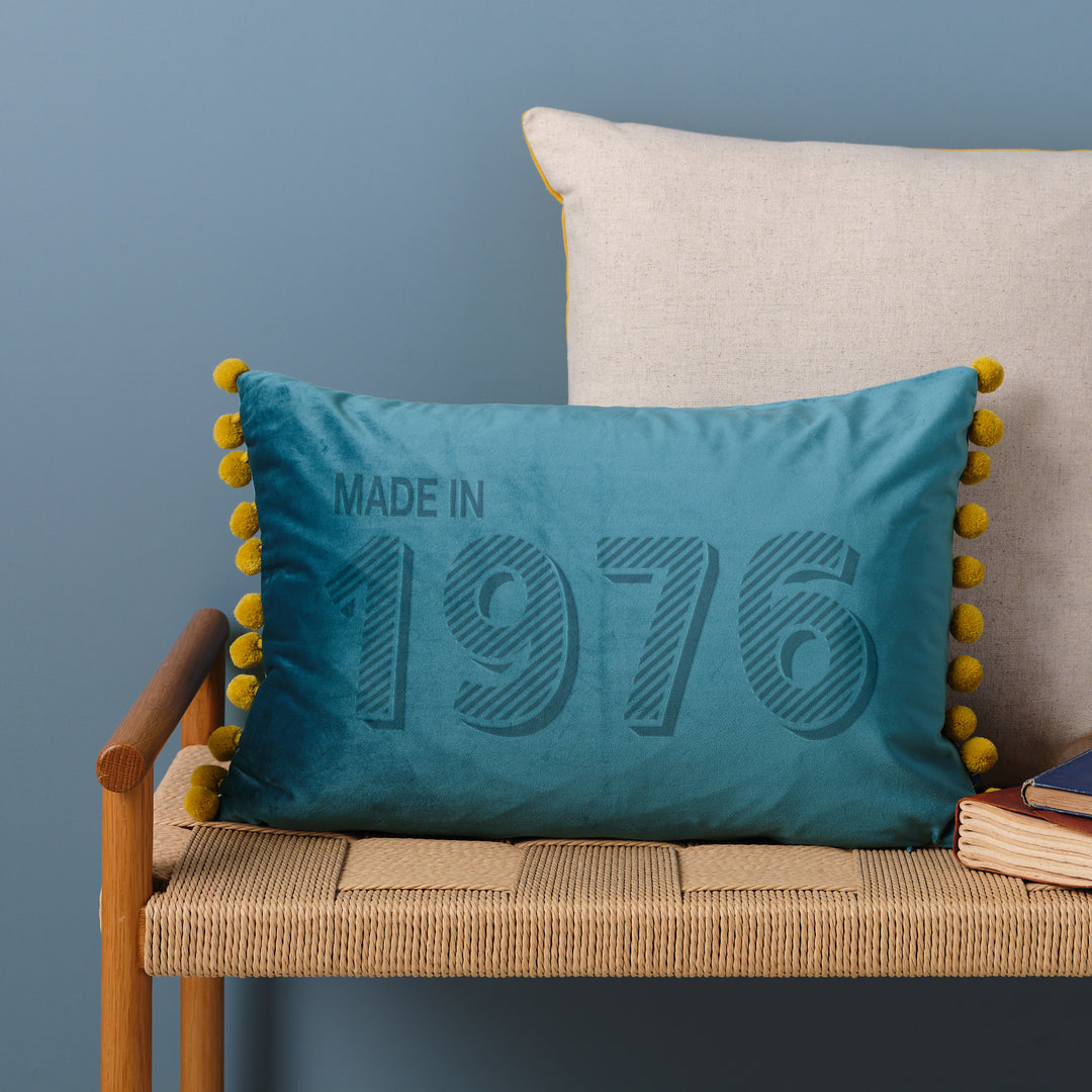A personalised 50th birthday gift velvet cushion with 'MADE IN 1976' laser engraved on it, placed on a wicker bench with other cushions and a book.