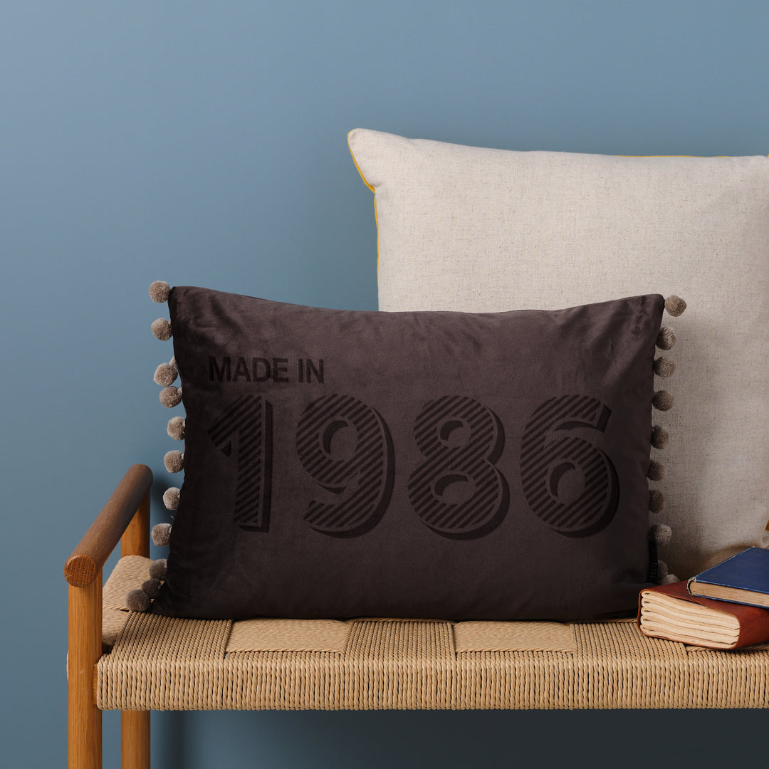 A personalised 40th birthday gift velvet cushion with 'MADE IN 1986' laser engraved on it, placed on a wicker bench with other cushions and a book.