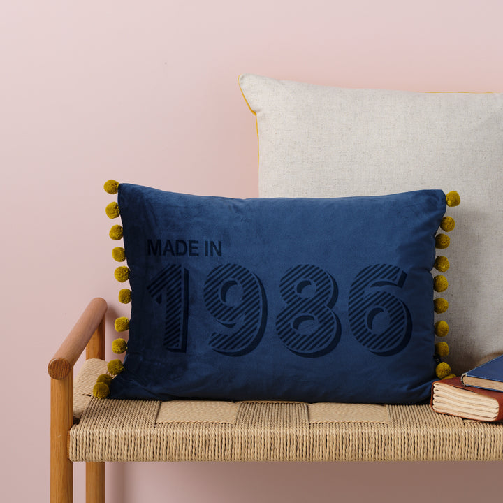 A personalised 40th birthday gift velvet cushion with 'MADE IN 1986' laser engraved on it, placed on a wicker bench with other cushions and a book.
